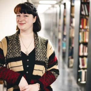 Olivia Mathers stands in the stacks and poses for a photo. She wears a stripped cardigan.
