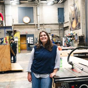 Avery McGarry stands in the scene shop wearing a blue sweater vest and smiling at the camera.