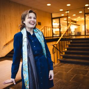Liz Barrett stands in front of steps wearing a dark blue dress and looking at the camera.
